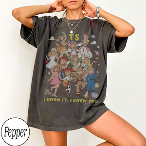 K&ouml;nnte beinhalten: Ein anthrazitfarbenes Kurzarm-T-Shirt mit einem Grafikdruck von mehreren animierten Figuren in verschiedenen Outfits. Der Text TS ist oben aufgedruckt und der Text i KNEW IT, i KNEW YOU ist unten auf dem Grafikdesign zu sehen.