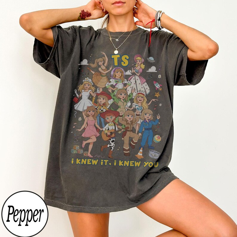 K&ouml;nnte beinhalten: Ein anthrazitfarbenes Kurzarm-T-Shirt mit einem Grafikdruck von mehreren animierten Figuren in verschiedenen Outfits. Der Text TS ist oben aufgedruckt und der Text i KNEW IT, i KNEW YOU ist unten auf dem Grafikdesign zu sehen.
