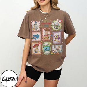 May include: A brown short-sleeve t-shirt featuring a graphic print of various cartoon characters in postage stamp-style frames. The central text reads "You've Got A Friend in Me" and "Hi!" with colourful illustrations of toys and space-themed elements.