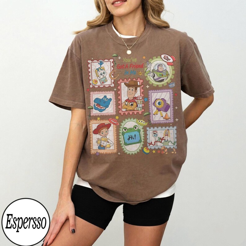 May include: A brown short-sleeve t-shirt featuring a graphic print of various cartoon characters in postage stamp-style frames. The central text reads "You've Got A Friend in Me" and "Hi!" with colourful illustrations of toys and space-themed elements.