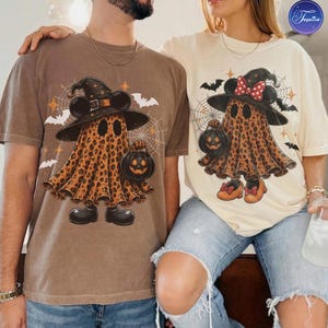 May include: A brown t-shirt featuring a leopard print ghost wearing a witch hat and holding a jack-o'-lantern, and a cream t-shirt featuring a leopard print ghost with a red polka dot bow, a witch hat, and a jack-o'-lantern, both with spider webs.
