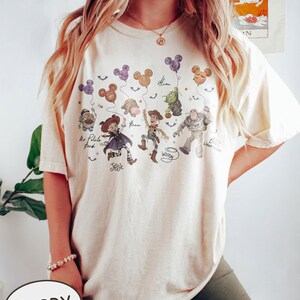 May include: Ivory-colored t-shirt featuring a graphic of Disney's Toy Story characters holding Mickey Mouse-shaped balloons. The characters include Mr. Potato Head, Jessie, Woody, Buzz Lightyear, and an Alien. The word "Ivory" is printed on the bottom.