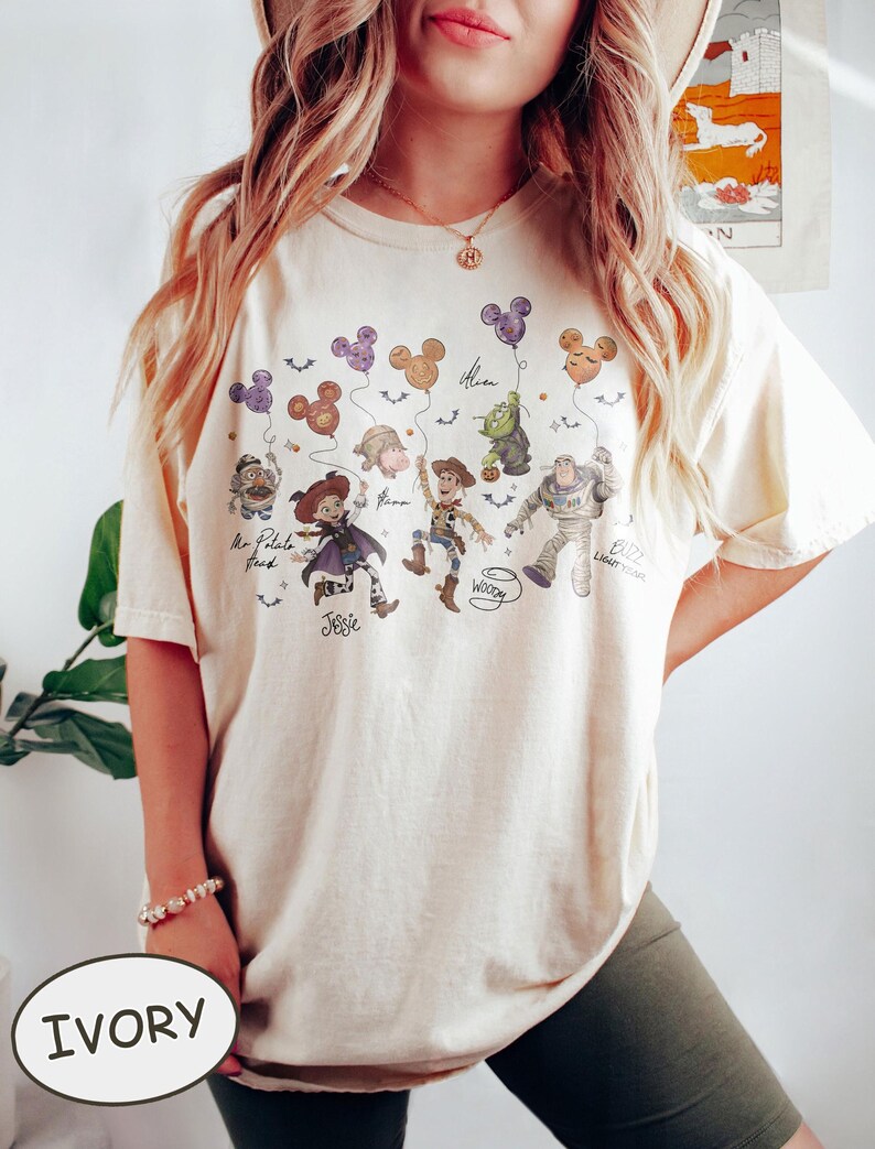 May include: Ivory-colored t-shirt featuring a graphic of Disney's Toy Story characters holding Mickey Mouse-shaped balloons. The characters include Mr. Potato Head, Jessie, Woody, Buzz Lightyear, and an Alien. The word "Ivory" is printed on the bottom.