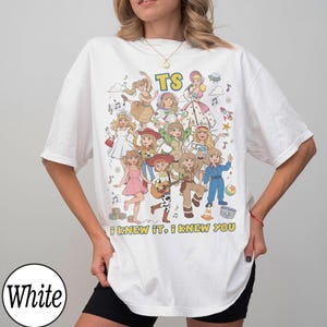 K&ouml;nnte beinhalten: Ein wei&szlig;es T-Shirt mit einem Grafikdruck von mehreren illustrierten Figuren in verschiedenen Outfits, darunter ein Raumanzug und Cowgirl-Kleidung. Der Text "TS" steht oben und der Schriftzug "I KNEW IT, I KNEW YOU" ist unten aufgedruckt.
