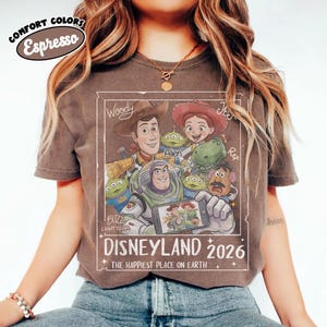 May include: Espresso-colored t-shirt featuring a graphic of Disney's Toy Story characters, including Woody, Buzz Lightyear, and Jessie. The design includes the text "DISNEYLAND 2026" and "THE HAPPIEST PLACE ON EARTH."