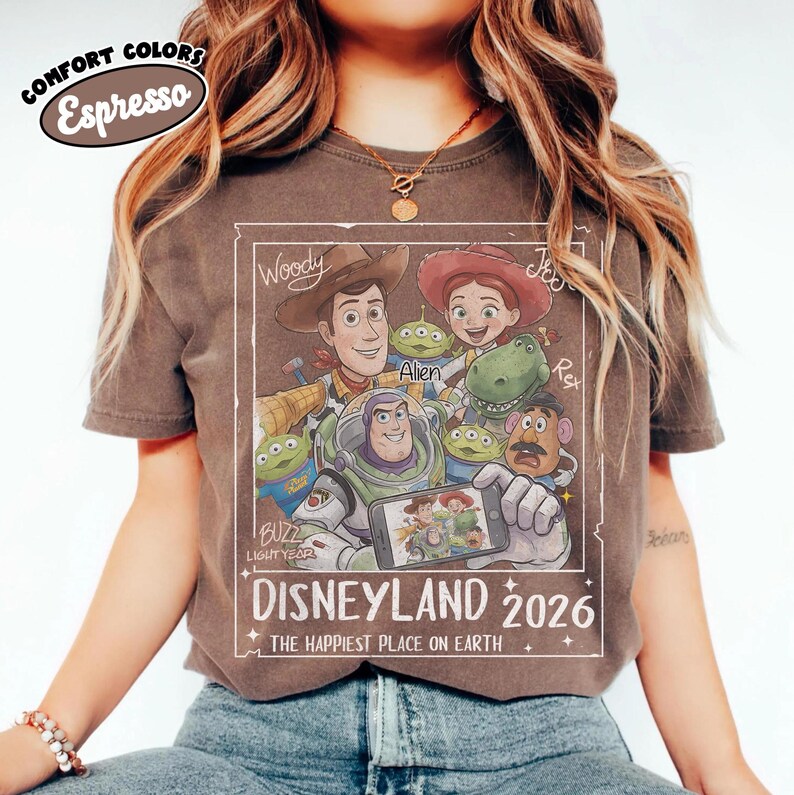 May include: Espresso-colored t-shirt featuring a graphic of Disney's Toy Story characters, including Woody, Buzz Lightyear, and Jessie. The design includes the text "DISNEYLAND 2026" and "THE HAPPIEST PLACE ON EARTH."
