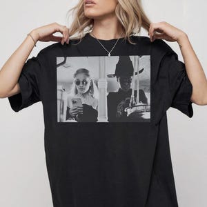 May include: Black t-shirt featuring a black and white image of two people, one wearing sunglasses and the other a hat, both looking at their phones. The shirt is a crew neck style.