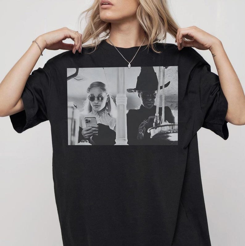 May include: Black t-shirt featuring a black and white image of two people, one wearing sunglasses and the other a hat, both looking at their phones. The shirt is a crew neck style.