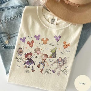 May include: Ivory-colored crew neck sweatshirt featuring a colorful graphic of Disney characters holding Mickey Mouse-shaped balloons. The design includes characters like Woody, Buzz Lightyear, and Jessie. The sweatshirt is made of a soft material.