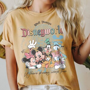 Puede incluir: Camiseta de color amarillo mostaza con un gr&aacute;fico de personajes de Disney, incluyendo Mickey Mouse, Minnie Mouse, Donald Duck y Goofy. La camiseta tiene el texto "Walt Disney's Disneyworld" y "Where dreams come true."
