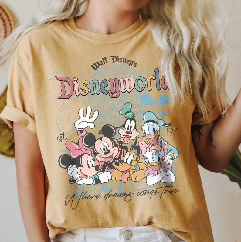 Puede incluir: Camiseta de color amarillo mostaza con un gr&aacute;fico de personajes de Disney, incluyendo Mickey Mouse, Minnie Mouse, Donald Duck y Goofy. La camiseta tiene el texto "Walt Disney's Disneyworld" y "Where dreams come true."