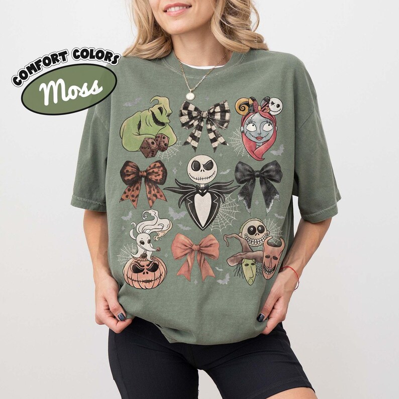 May include: A moss green short-sleeve t-shirt featuring a graphic print of characters and bows from The Nightmare Before Christmas. The design includes Jack Skellington, Sally, Zero, Oogie Boogie, and various decorative bows on a muted background.