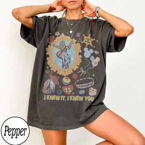 May include: A charcoal gray short-sleeve t-shirt featuring a graphic print with a central ornate gold frame containing a figure with a guitar. Additional graphics include a sheriff star with text Infinity and Beyond, balloons, a cowboy boot, a ball, and the text I KNEW IT, I KNEW YOU.