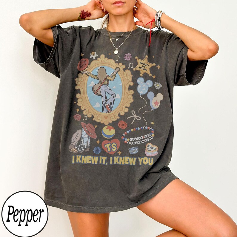 May include: A charcoal gray short-sleeve t-shirt featuring a graphic print with a central ornate gold frame containing a figure with a guitar. Additional graphics include a sheriff star with text Infinity and Beyond, balloons, a cowboy boot, a ball, and the text I KNEW IT, I KNEW YOU.