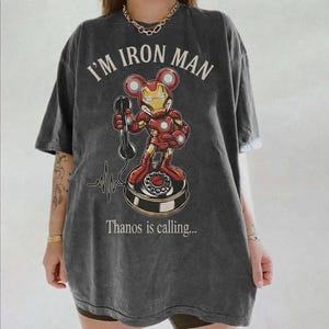 Op de afbeelding: Donkergrijs t-shirt met een cartoon Iron Man-personage met Mickey Mouse-oren die een telefoon vasthoudt. De tekst "I'M IRON MAN" staat boven het personage, met "Thanos is calling..." eronder. Er is ook een hartslaglijn.