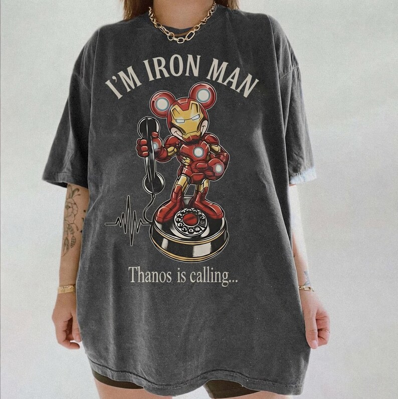 Op de afbeelding: Donkergrijs t-shirt met een cartoon Iron Man-personage met Mickey Mouse-oren die een telefoon vasthoudt. De tekst "I'M IRON MAN" staat boven het personage, met "Thanos is calling..." eronder. Er is ook een hartslaglijn.