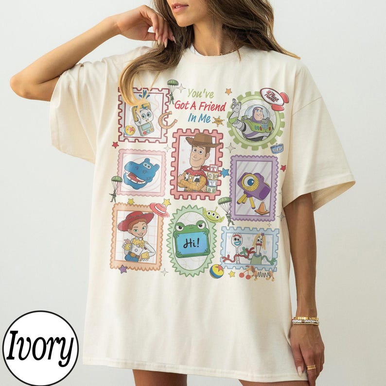 May include: An ivory short-sleeve t-shirt featuring a colourful graphic print of various animated characters from Toy Story arranged in postage stamp-style frames. The text "You've Got A Friend In Me" is printed in the centre among the characters.