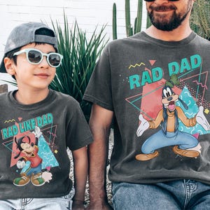 May include: Matching charcoal gray t-shirts with the text "RAD DAD" and "RAD LIKE DAD" in teal, pink, and yellow. The shirts feature a cartoon Goofy graphic. A child wears sunglasses and a gray cap.
