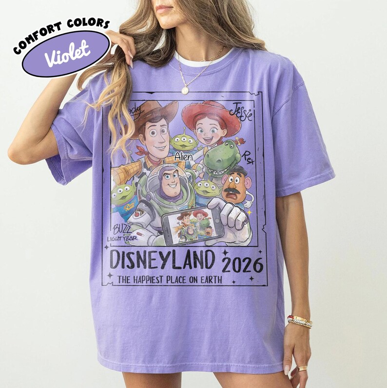 May include: A violet Comfort Colors t-shirt featuring a graphic of Toy Story characters, including Woody, Buzz Lightyear, and Jessie. The design includes the text "DISNEYLAND 2026" and "THE HAPPIEST PLACE ON EARTH."