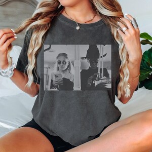 May include: Dark gray t-shirt featuring a black and white image of two people. One person is wearing sunglasses and holding a phone, and the other is wearing a hat and holding an object. The shirt is a casual style.