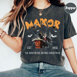 May include: Dark gray t-shirt with an orange "MANOR" graphic. The design features characters and imagery from "The Nightmare Before Christmas," including bats, a spiderweb, and a jack-o'-lantern. The text "EST. 1973" and "THE NIGHTMARE BEFORE CHRISTMAS" are also included.