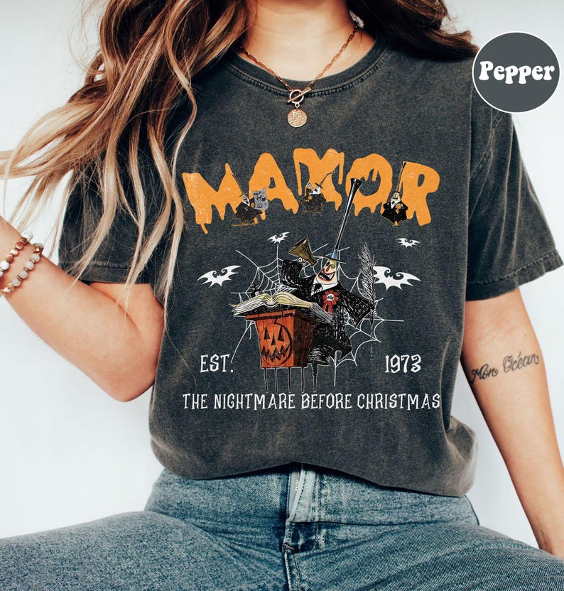 May include: Dark gray t-shirt with an orange "MANOR" graphic. The design features characters and imagery from "The Nightmare Before Christmas," including bats, a spiderweb, and a jack-o'-lantern. The text "EST. 1973" and "THE NIGHTMARE BEFORE CHRISTMAS" are also included.