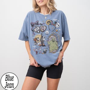 Może przedstawiać: Niebieska koszulka w kolorze jeansu z nadrukiem graficznym przedstawiającym postacie z Miasteczka Halloween, w tym Jacka Skellingtona, Sally, Oogie Boogie i Zero, wraz z tematycznymi ikonami takimi jak mysie uszy, kokardy i dynie.