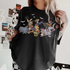 May include: Black t-shirt featuring a Halloween-themed graphic of Winnie the Pooh and friends trick-or-treating. The design includes bats, spiderwebs, and characters holding pumpkin buckets. The shirt is a dark color and has a vintage look.