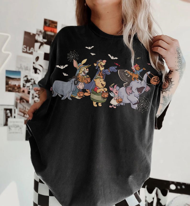 May include: Black t-shirt featuring a Halloween-themed graphic of Winnie the Pooh and friends trick-or-treating. The design includes bats, spiderwebs, and characters holding pumpkin buckets. The shirt is a dark color and has a vintage look.