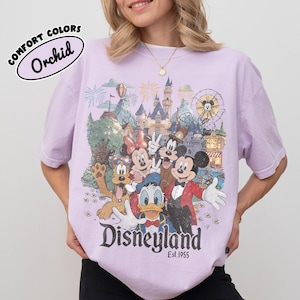 May include: Orchid-colored t-shirt featuring a Disneyland graphic with Mickey Mouse, Minnie Mouse, Donald Duck, Goofy, and other characters. The graphic includes a castle, fireworks, and a Ferris wheel. The text "Disneyland Est. 1955" is printed below the characters.