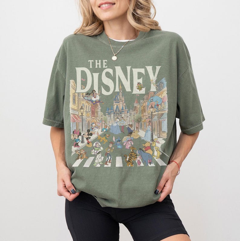 May include: An olive green short-sleeve t-shirt featuring a large graphic print with the text THE DISNEY in a cream font. The illustration depicts a street scene with a castle in the background and various animated characters walking across a crosswalk.