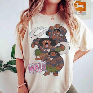 K&ouml;nnte beinhalten: Ein cremefarbenes T-Shirt mit einem Grafikdruck der Figur Maui aus dem Film Moana. Das Design zeigt drei Versionen der Figur in verschiedenen Posen mit Stammes-Tattoos, die einen gro&szlig;en Haken halten, sowie den Text MAUI in Pink.