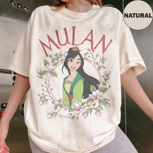 May include: A natural-colored t-shirt featuring a graphic of Mulan in a green and yellow outfit, surrounded by a circular frame of pink magnolia flowers. The word MULAN is printed in large red letters above the illustration of the character.
