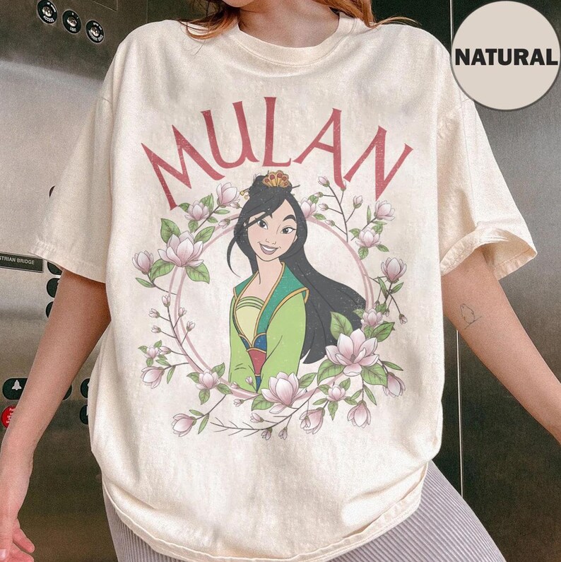 May include: A natural-colored t-shirt featuring a graphic of Mulan in a green and yellow outfit, surrounded by a circular frame of pink magnolia flowers. The word MULAN is printed in large red letters above the illustration of the character.