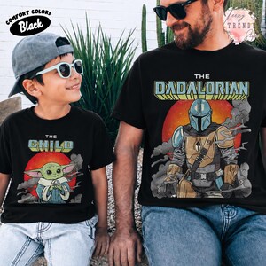 May include: Two black t-shirts featuring graphic prints. The smaller shirt displays a green creature with the text THE CHILD above a red sun. The larger shirt shows a armored character with the text THE DADALORIAN above a red sun.