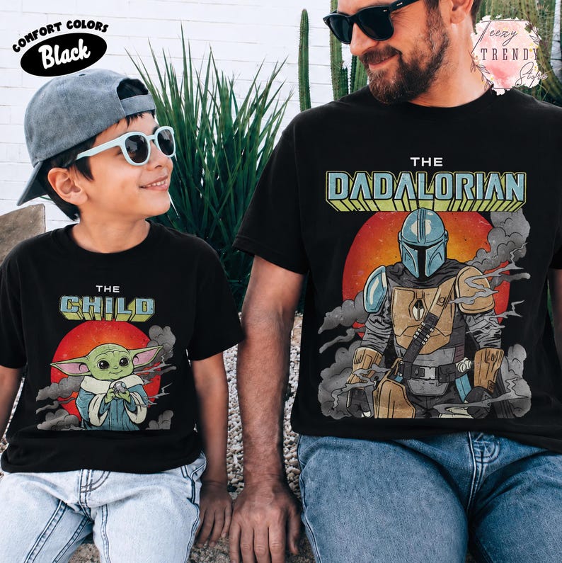 May include: Two black t-shirts featuring graphic prints. The smaller shirt displays a green creature with the text THE CHILD above a red sun. The larger shirt shows a armored character with the text THE DADALORIAN above a red sun.