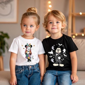 May include: Two young children wearing graphic t-shirts. One child wears a white tee with a Minnie Mouse design in fall colors. The other child wears a black tee with a Jack Skellington Mickey Mouse design, bats, and ghosts.