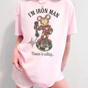 Op de afbeelding: Lichtroze T-shirt met de tekst "I'M IRON MAN" boven een cartoonillustratie van Iron Man als Mickey Mouse op een draaitelefoon. De tekst "Thanos is calling..." staat eronder.
