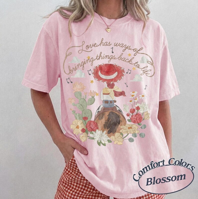 May include: A light pink short-sleeve t-shirt featuring a graphic of a cowgirl hat, a cactus, and flowers. The text reads, Love has ways of bringing things back to life. The design includes musical notes and clouds in a soft, watercolor-style illustration.