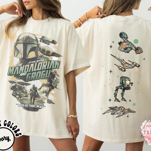 Pode incluir: Uma t-shirt cor de marfim com um gr&aacute;fico frontal de The Mandalorian e Grogu com o texto "The Mandalorian and Grogu" e "This is the way". O gr&aacute;fico traseiro mostra quatro ve&iacute;culos e droides de Star Wars dispostos verticalmente com pequenas estrelas.