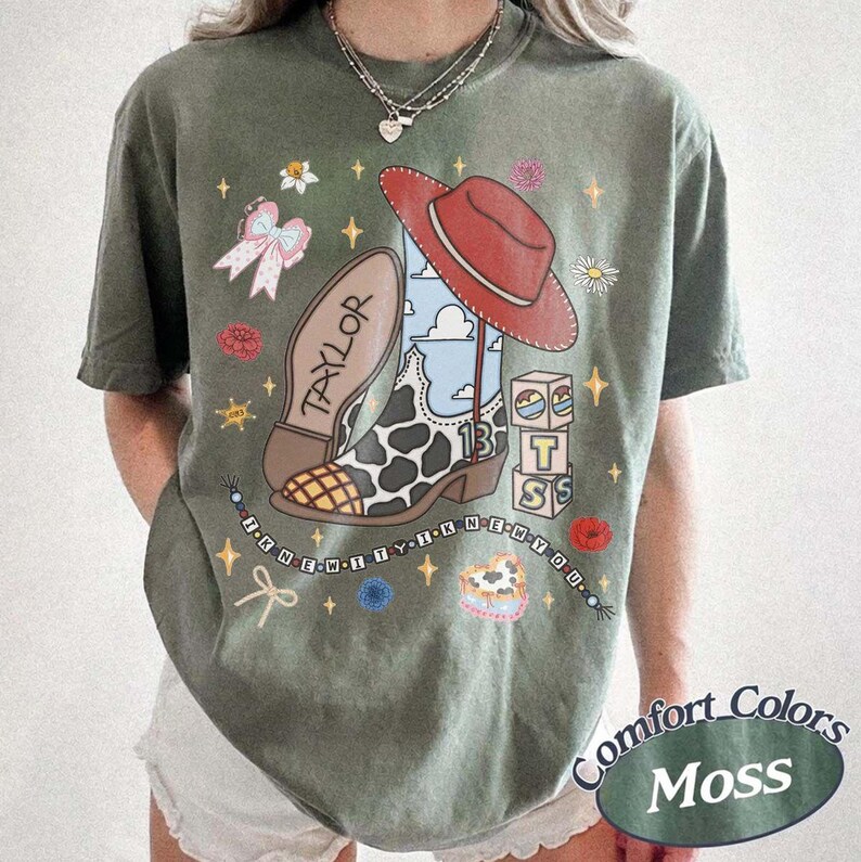 May include: A moss green short-sleeve t-shirt featuring a graphic print of a cow-print cowboy boot with a red hat, the name TAYLOR, the number 13, stacked blocks, a bow, and the text I KNEW YOU WERE TROUBLE in a beaded necklace design.