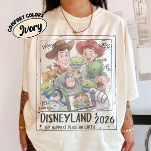 May include: Ivory-colored t-shirt featuring a graphic of Disney's Toy Story characters. The design includes Woody, Jessie, Buzz Lightyear, and aliens, with the text "DISNEYLAND 2026" and "THE HAPPIEST PLACE ON EARTH".