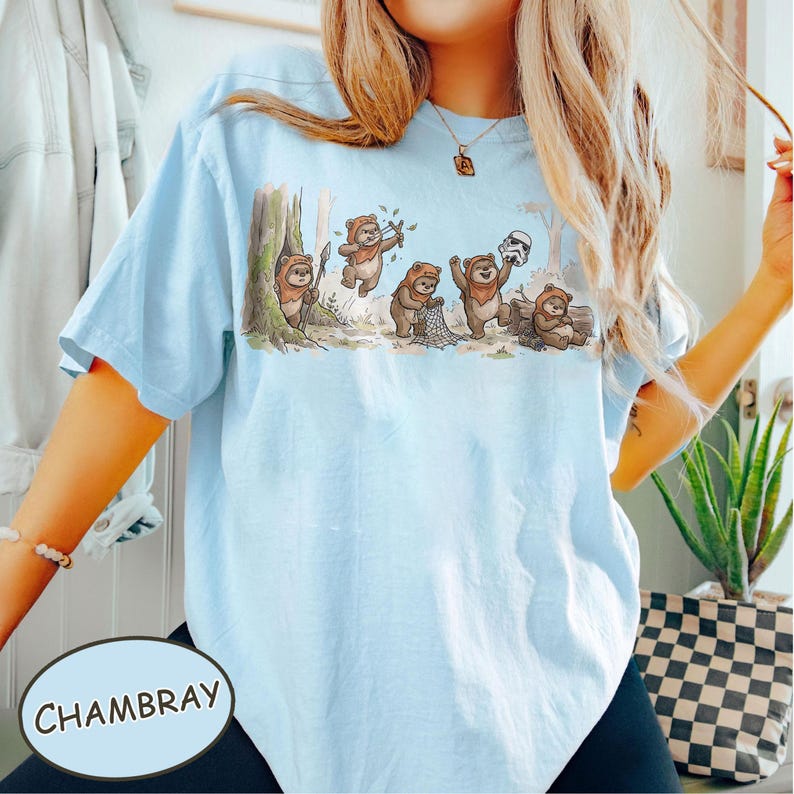 May include: Light blue t-shirt with a graphic of cartoon Ewoks in a forest setting. The Ewoks are depicted in various poses, including holding weapons and a Stormtrooper helmet. The word "Chambray" is printed on the bottom left.