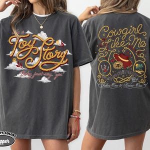 Może przedstawiać: Ciemnoszary t-shirt z kr&oacute;tkim rękawem z grafiką z przodu z napisem Toy Story i chmurami oraz grafiką z tyłu z napisem Cowgirl like me, Takes One to Know One, kapeluszem kowbojskim, podkową i elementami dekoracyjnymi w kształcie liny.