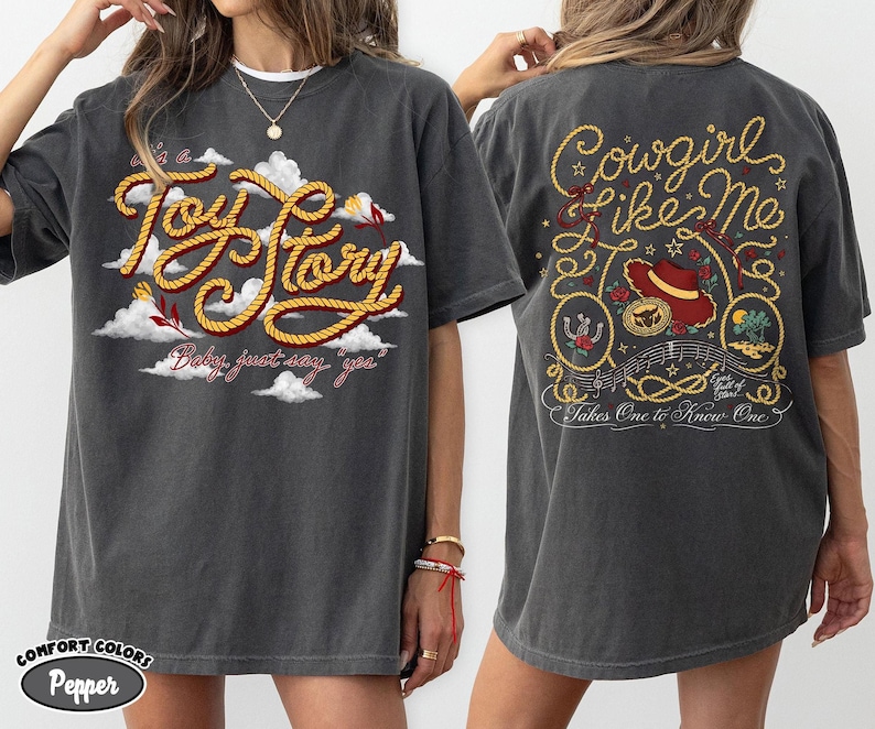 Może przedstawiać: Ciemnoszary t-shirt z kr&oacute;tkim rękawem z grafiką z przodu z napisem Toy Story i chmurami oraz grafiką z tyłu z napisem Cowgirl like me, Takes One to Know One, kapeluszem kowbojskim, podkową i elementami dekoracyjnymi w kształcie liny.