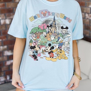 May include: Light blue t-shirt featuring a colorful graphic of Disney characters and the text "Walt Disney World." The design includes Mickey Mouse, Minnie Mouse, Donald Duck, and other characters in a cartoon style.