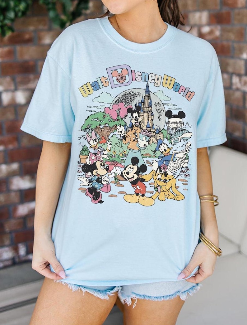 May include: Light blue t-shirt featuring a colorful graphic of Disney characters and the text "Walt Disney World." The design includes Mickey Mouse, Minnie Mouse, Donald Duck, and other characters in a cartoon style.