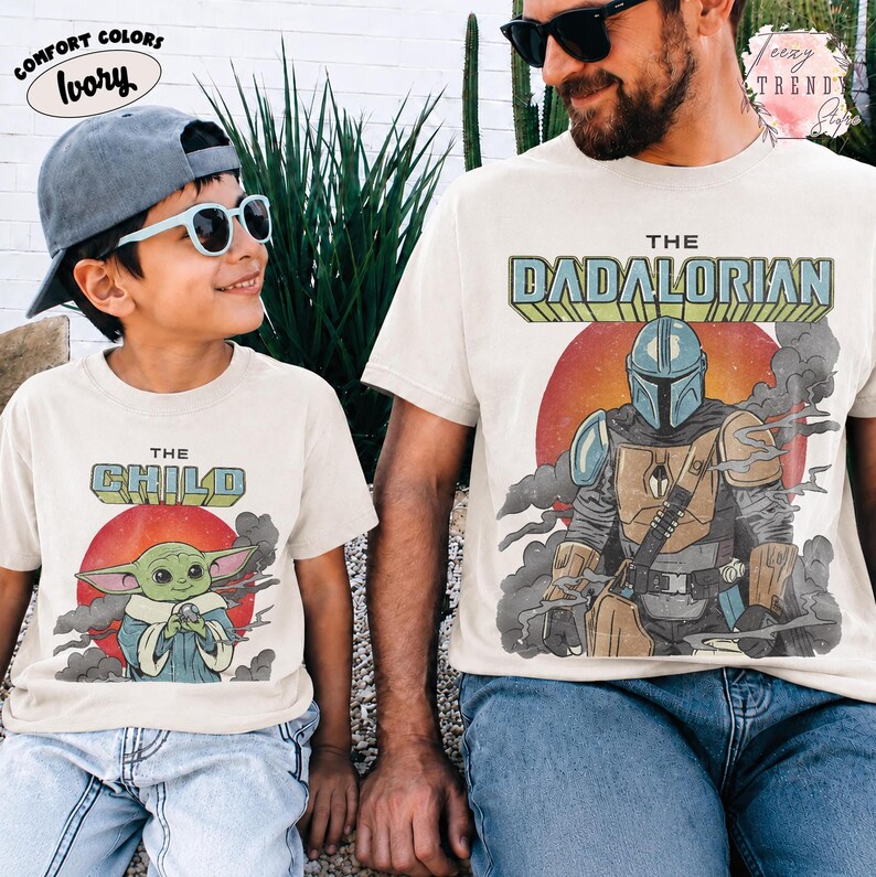 May include: Two ivory t-shirts featuring graphic prints. The smaller shirt displays a green character with large ears and the text THE CHILD. The larger shirt shows a helmeted figure in armor with the text THE DADALORIAN. Both have red suns.
