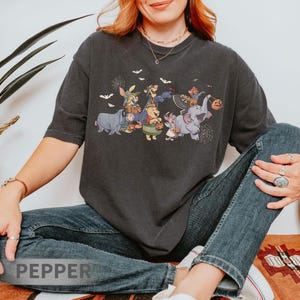 May include: Dark gray t-shirt featuring a Halloween-themed graphic of cartoon characters, including Winnie the Pooh and friends, dressed in costumes. Bats and spiderwebs are also included in the design. The shirt is worn by a person.