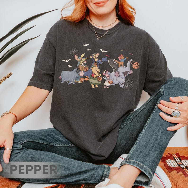 May include: Dark gray t-shirt featuring a Halloween-themed graphic of cartoon characters, including Winnie the Pooh and friends, dressed in costumes. Bats and spiderwebs are also included in the design. The shirt is worn by a person.